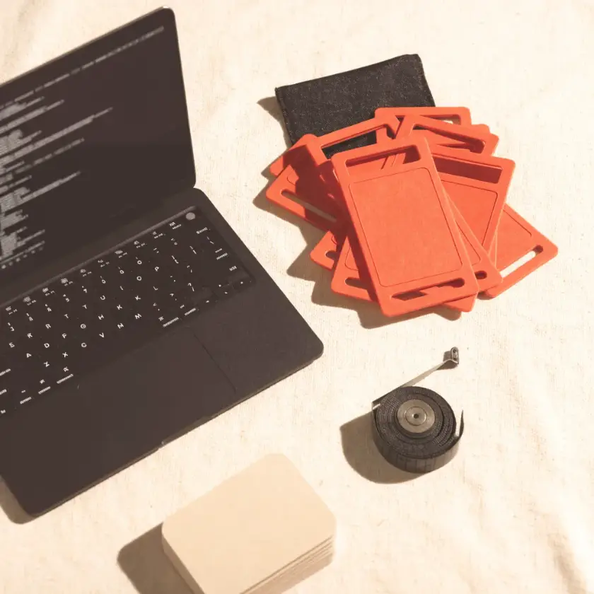 Banc de lucru văzut de sus: laptop cu cod, zip-tie-uri coral și etichete goale pe bumbac crem